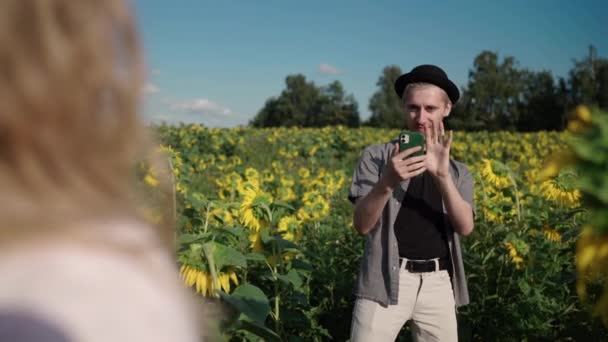 Blond flicka i ett solrosfält läser bok, poserar på telefonen till söt kille — Stockvideo