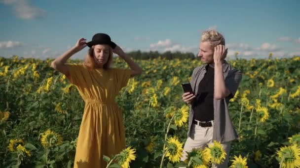 Red-haired women on sunflower field is photographed on the phone guy in hat — Stock Video