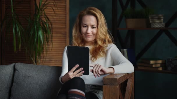 Женщина с рыжими волосами читает книгу на планшете в мансарде, улыбается, думает — стоковое видео