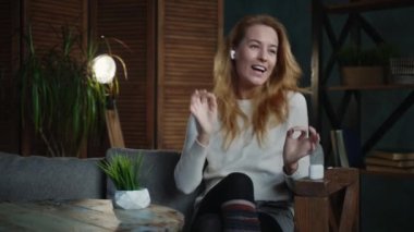 Kızıl saçlı beyaz kadın tavan arası odasında kanepenin üzerinde podcast dinliyor.