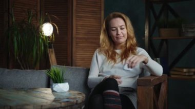 Kızıl saçlı beyaz kadın tavan arası odasında kanepenin üzerinde podcast dinliyor.