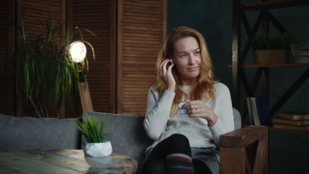 Donna caucasica dai capelli rossi in camera in stile loft sul divano ad ascoltare podcast — Video Stock