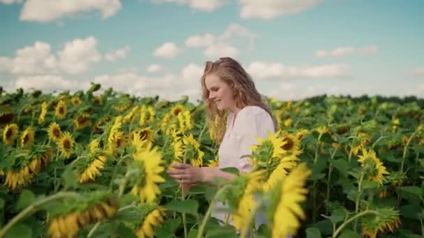 Молодая блондинка-кавказка с вьющимися волосами гуляет по полю подсолнухов улыбается — стоковое видео