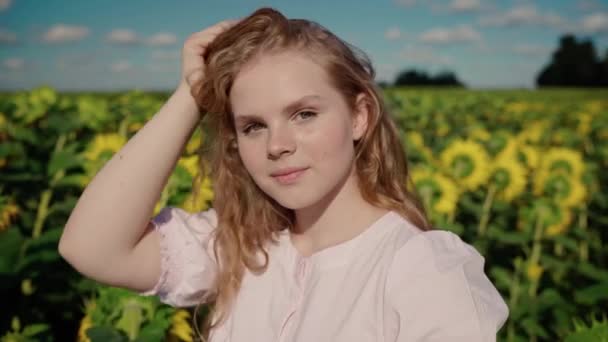 Junge blonde kaukasische Mädchen in Sonnenblumenfeld lächelt, glätten lockiges Haar — Stockvideo