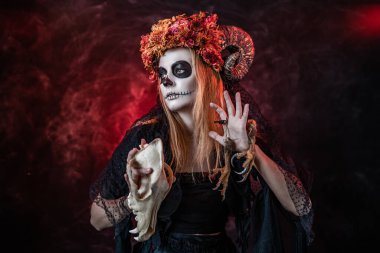 Boynuzları ve kafasında kurumuş çiçekleri olan cadı kafatası, bacakları tutan Faunus gibi.