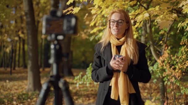Operator registreert videoreportage met roodharige vrouw in bril in herfstpark — Stockvideo
