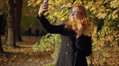 Kadın sonbahar ormanında yürüyor, bir buket yaprakla fotoğraf çekiyor, gülümsüyor.