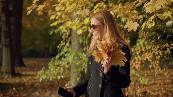 Kobieta spaceruje po jesiennym lesie, robi zdjęcia bukietem liści, uśmiecha się — Wideo stockowe