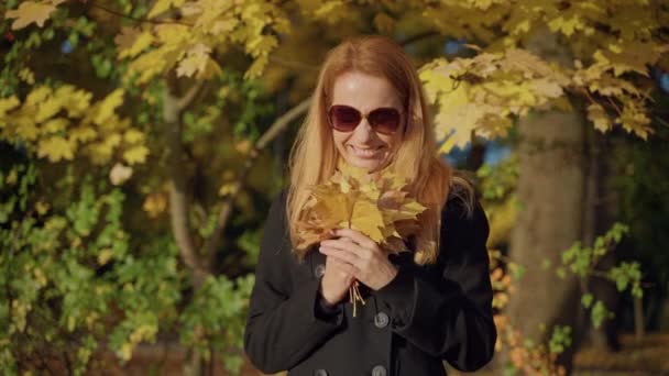 Wanita berambut merah berjalan di taman musim gugur dengan kacamata hitam, mengagumi buket daun — Stok Video