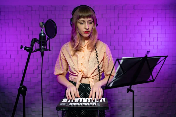 Chica en auriculares mira en el puesto de música, toca un teclado en un estudio brillante — Foto de Stock