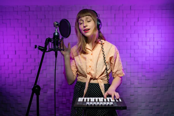 Chica en auriculares con los ojos cerrados canta en el micrófono, toca el teclado — Foto de Stock