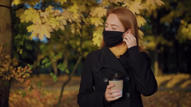 Kobieta zdejmuje maskę ochronną, pije kawę z kubka rzemiosła w jesiennym parku — Wideo stockowe