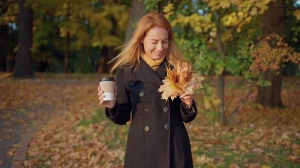 Woman walks in autumn forest, with bouquet of maple leaves, craft paper cup — Stock Video