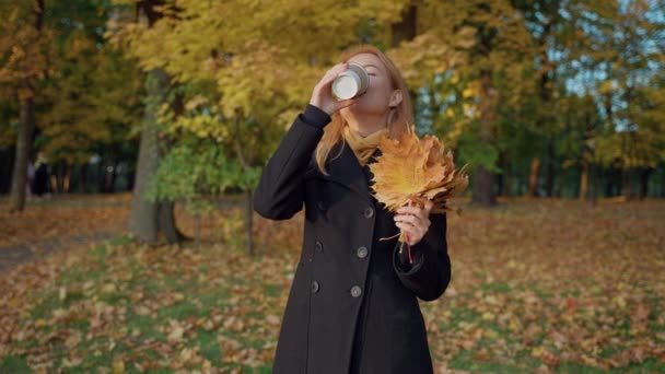Γυναίκα στο πάρκο φθινόπωρο, με το τσάι σε ένα κύπελλο χειροτεχνίας, πετά μπουκέτο φύλλα, χαμόγελα — Αρχείο Βίντεο