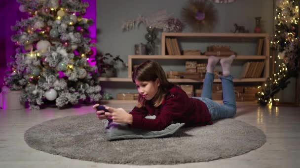 Έφηβος τύπος ξαπλωμένος στο χαλί παρακολουθεί βίντεο στο τηλέφωνο την παραμονή της Πρωτοχρονιάς στο σαλόνι — Αρχείο Βίντεο