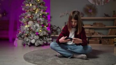 Genç bir çocuk Noel arifesinde halıda oturmuş akıllı bir telefonla oyun oynuyor.