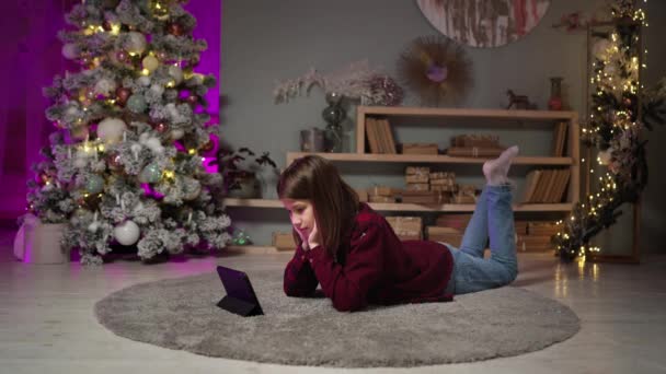 Adolescente se encuentra en la alfombra, lee el libro electrónico en la víspera de Año Nuevo. Esperando la celebración — Vídeo de stock