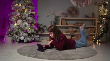 Uzun saçlı genç halıda yatıyor, yılbaşı arifesinde tablete bakıyor.