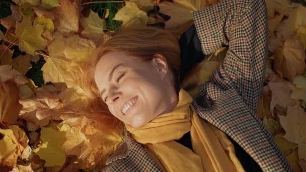 再生コートリラックスの赤い髪の若い女性は、黄色のカエデの葉に横たわっている — ストック動画