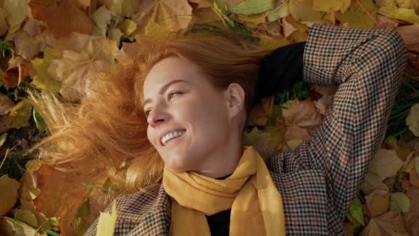 Meisje ligt op esdoorn herfst bladeren, zet palmen achter hoofd, kijkt in frame — Stockvideo