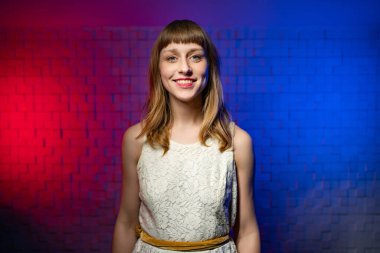 Kahverengi saçlı, beyaz elbiseli kız kameraya bakıyor, gülümsüyor.