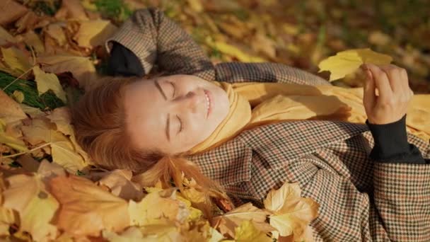 Female lying in autumn foliage in park, looks at sky, smiles, breathes fresh air — ストック動画