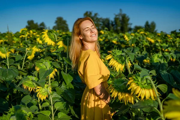 Beautiful red-haired woman in yellow dress enjoys fresh air in sunflower field Royalty Free Stock Photos