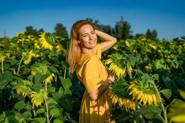 Red-haired lady in yellow dress smiling, squinting at sun in sunflower field Stock Picture