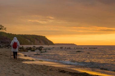 Kırmızı sırt çantalı beyaz ceketli bir adam akşam vakti deniz kıyısında İskandinav yürüyüşü yapıyor..