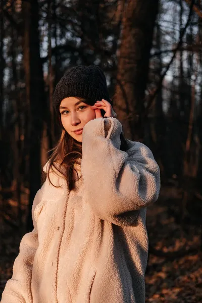 Smilende ung kvinde i frakke og vinter hat stående i efteråret skov. Kvinde kigger på kameraet på træers baggrund og indånder frisk luft. Person i naturen. Have det sjovt udendørs. - Stock-foto