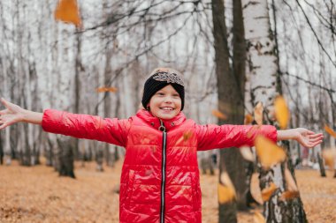 Ayakta gülen ve kuru yaprakları üzerine atan küçük bir kız. Mutlu çocuk sonbahar ormanında oynuyor. Gülümseyen çocuk dışarıda eğleniyor. Kırmızı ceket, örgü saç bandı..