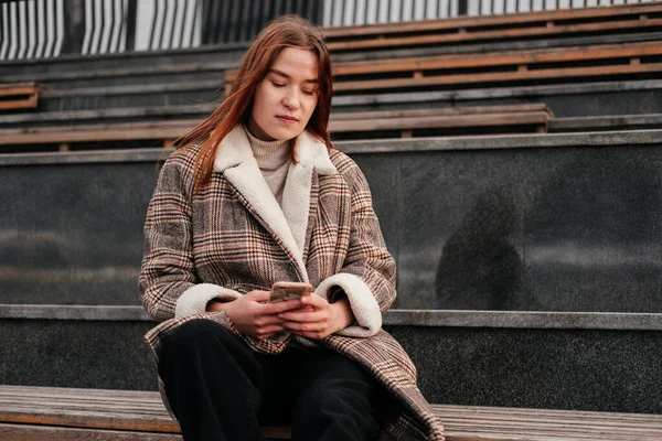 通りの木製のベンチに一人で座って手にスマートフォンでコートを着た若い女性 屋外でモバイルデバイスに取り組んでいます 女性フリーランスブロガー テキスト チャット — ストック写真