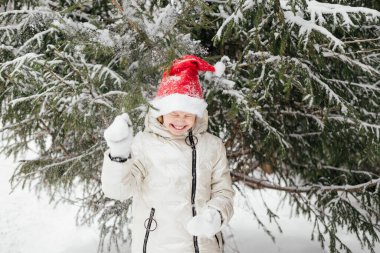 Kırmızı Noel şapkalı gülen küçük kız ormandaki köknar ağacının altında duruyor. Açık hava kış aktiviteleri. Noel Baba şapkalı mutlu çocuk oynuyor, kış ormanında eğleniyor..