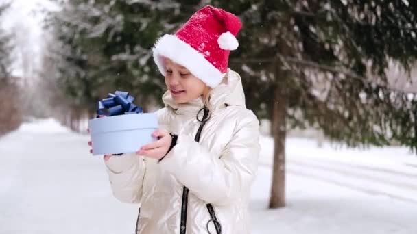 Улыбающаяся маленькая девочка в красной рождественской шляпе, трясущая синей подарочной коробкой. Счастливый ребенок в шляпе Санта наслаждается подарком в зимнем лесу. Зимние мероприятия на открытом воздухе. — стоковое видео