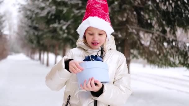 Маленькая девочка в красной рождественской шляпе открывает синюю подарочную коробку и удивляется, удивляется, шокируется. Счастливый ребенок в шляпе Санта наслаждается подарком в зимнем лесу. Зимние мероприятия на открытом воздухе. — стоковое видео