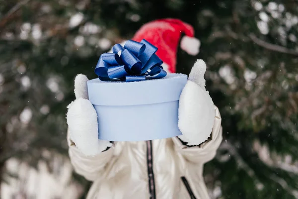 赤いクリスマスの帽子に青いギフトボックスを持ってる正体不明の少女。冬の森でプレゼントを楽しむサンタの帽子の幸せな子供。冬の野外活動. — ストック写真