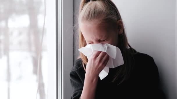 Ill child in sweater sitting on window sill at home. Sick little blonde girl sneezing into napkin. Covid-19 and symptoms after vaccine. Healthcare, medicine. Seasonal allergy, cold, runny nose — Stock Video