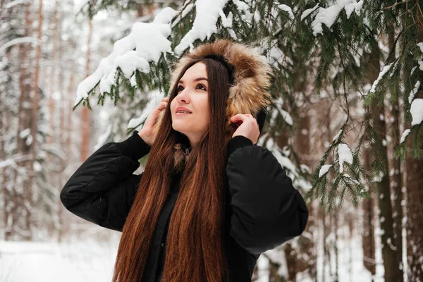 Smiling young woman in black coat holding hood standing under fir tree in wintre forest. Girl with long hair in park outdoors — Stock Photo, Image