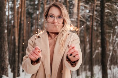 Açık havada kış ormanlarında elinde maytap tutan bej ceketli ve kazaklı orta yaşlı bir kadın. Gözlüklü sarışın kız.