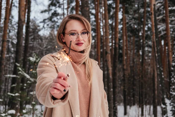 Blonde mid age woman in winter forest with sparkler in hand. Woman in coat and sweater outdoors. Girl in eyeglasses — Stock Photo, Image
