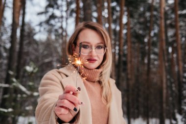 Kış ormanında elinde maytapla orta yaşlı bir kadın. Dışarıdaki kadın ceketli ve kazaklı. Gözlüklü sarışın kız.