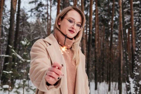 Blonde mid age woman in winter forest with sparkler in hand. Woman in coat and sweater outdoors. Girl in eyeglasses — Stock Photo, Image