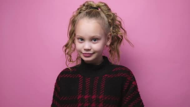 Little blonde girl in sweater makes heart symbol with hands and smiling on pink background. Valentines day love gesture. Caucasian child studio shot — Stock video