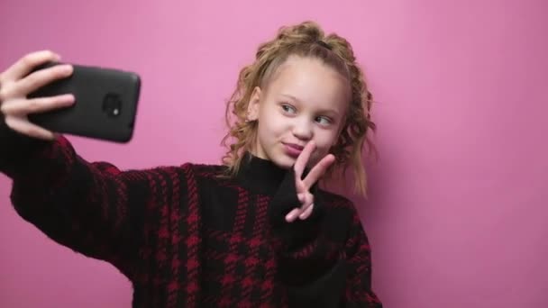 Blonde little girl in sweater taking selfie on pink background. Smiling child using smartphone. Internet and gadget kids addiction — Wideo stockowe