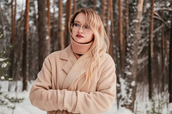 Frozen mid age woman in coat and sweater standing in winter forest warming up hands in sleeves outdoors. Blonde girl in eyeglasses — Stock Photo, Image