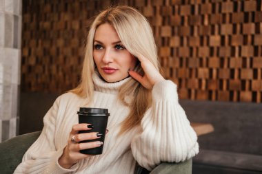 Orta yaşlı bir kadın kafede oturuyor ve gözlerini kaçırarak kahve içiyor. Beyaz süveter giyen beyaz sarışın kız..