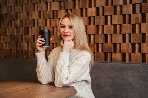 Smiling mid age woman holding cup of coffee sitting at cafe indoors. Caucasian girl in white sweater