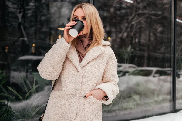 外を眺めながらコーヒーを飲む中年女性の笑顔 白人ブロンド女の子で白いコートオンストリート — ストック写真