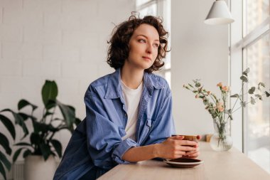 Kıvırcık saçlı, gülümseyen genç bir kadın kafede oturuyor ve içeride kahve içiyor.