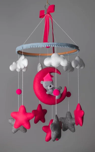 Baby crib hangings, Felt cot mobile with stuffed soft stars clouds and moon with sleeping teddy, Cot isolated against neutral grey color background. A cute teddy wearing a pink hat. Baby room decor.